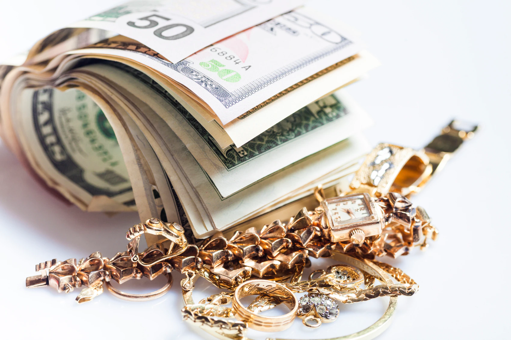 Rolled stack of US dollar bills beside gold jewelry, including rings, bracelets, and a watch.