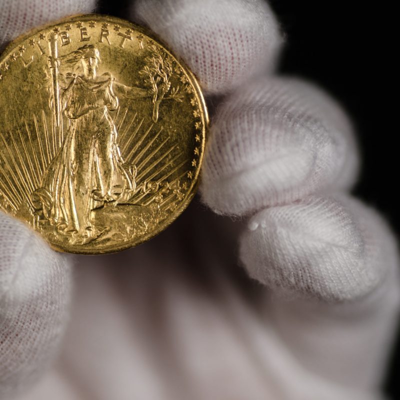 A white-gloved hand holding a gold coin with a depiction of Lady Liberty on the front.