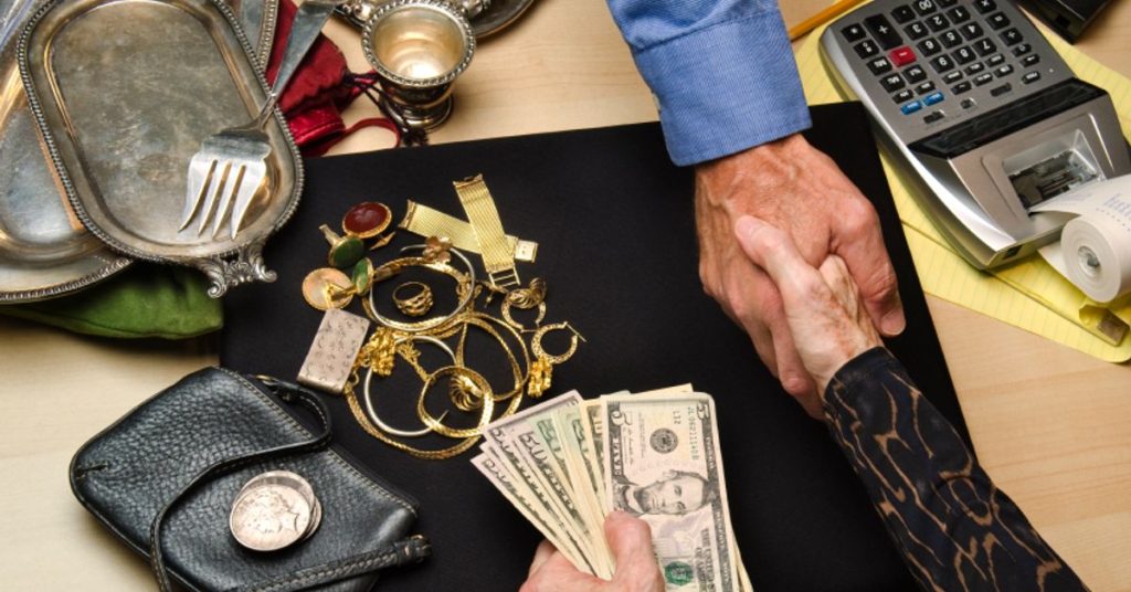 Handshake over a table with jewelry, silver items, and cash during a pawnshop transaction