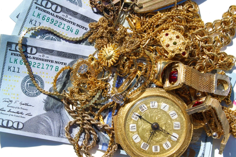 Gold jewelry and vintage watch placed over hundred‑dollar bills symbolizing cash for gold
