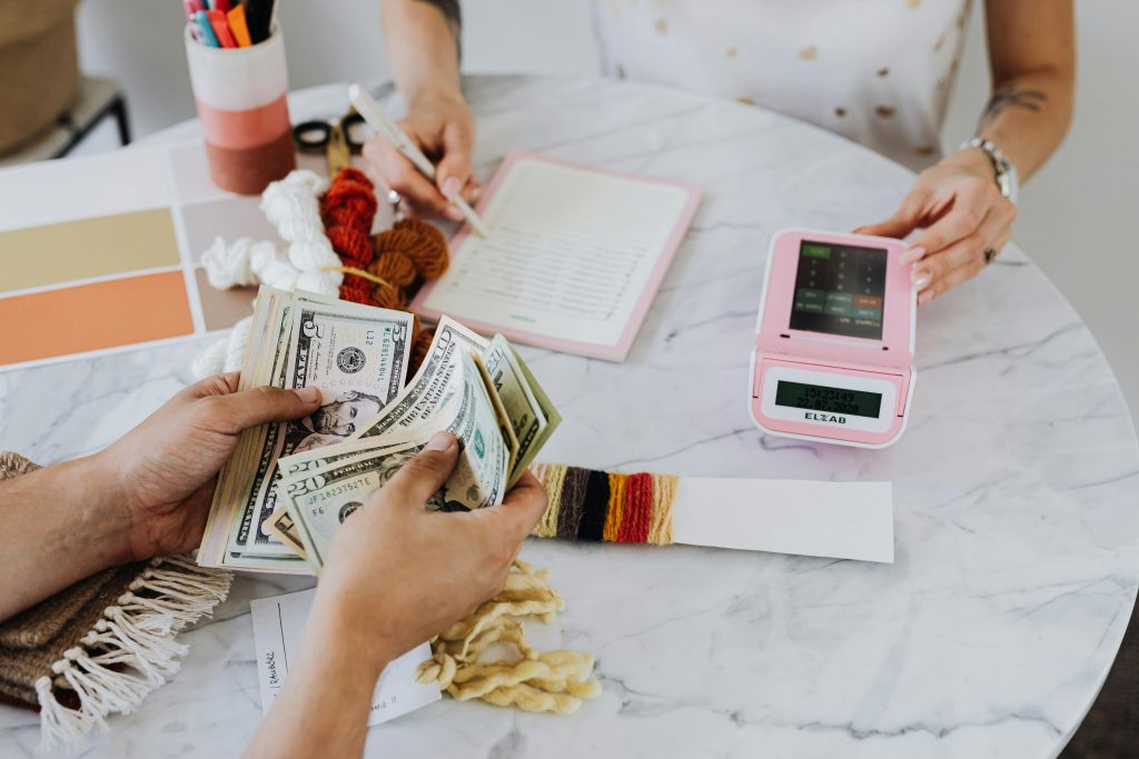 Two people exchanging U.S. dollar bills and recording details at a table with colorful yarn samples